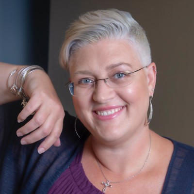 A person with short, silver hair and glasses is smiling at the camera. They are wearing a purple top and a dark cardigan, accessorized with large hoop earrings, a cross necklace, and several bracelets. One arm is relaxed over the back of a chair.