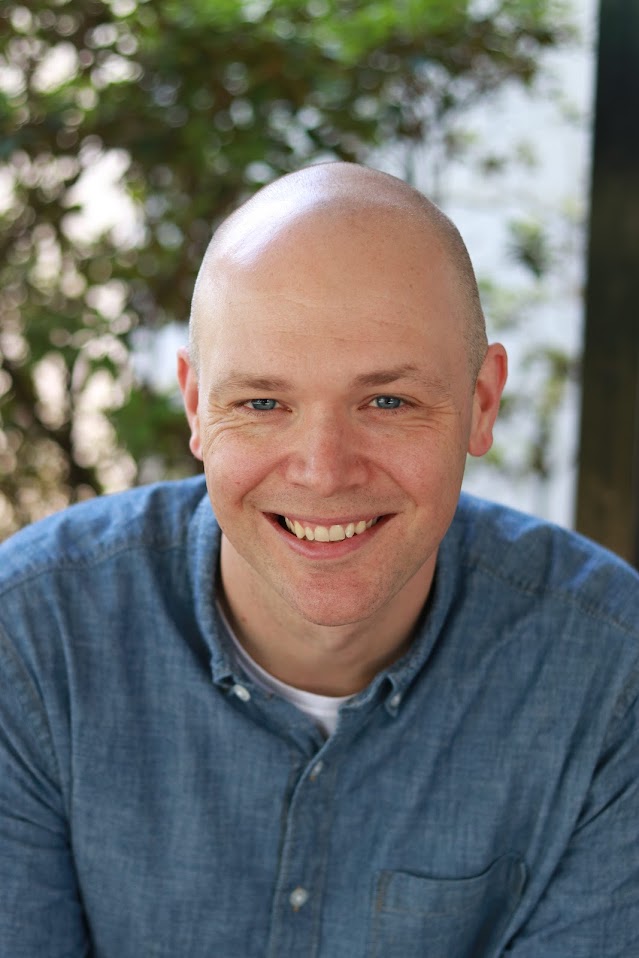 A bald man in a denim shirt smiles at the camera with greenery in the background.