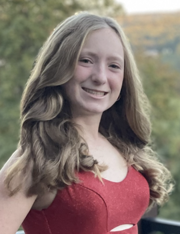 A young woman with long wavy hair smiles while wearing a red dress. She stands outside with trees and a blurred landscape in the background.