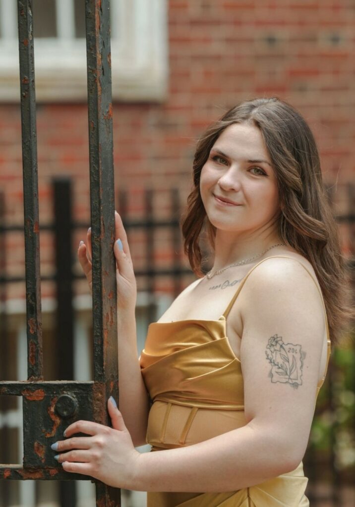 A person with long brown hair in a gold dress stands outdoors near a metal gate, holding onto it with one hand. A tattoo is visible on their right arm. Brick wall in the background.
