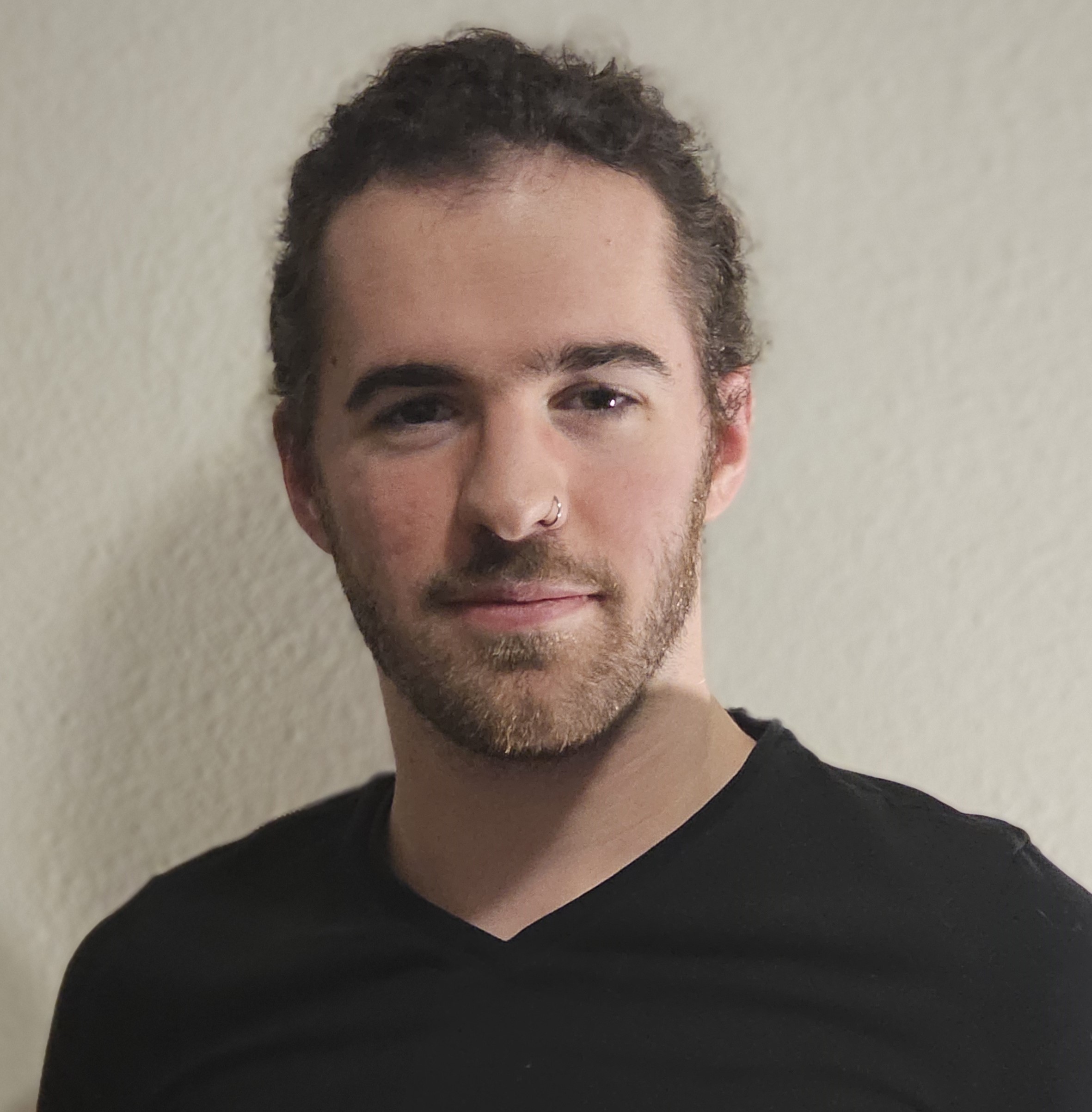 A person with a beard and long hair tied back is wearing a black V-neck shirt. They have a neutral expression and a nose ring, standing against a plain light-colored background.