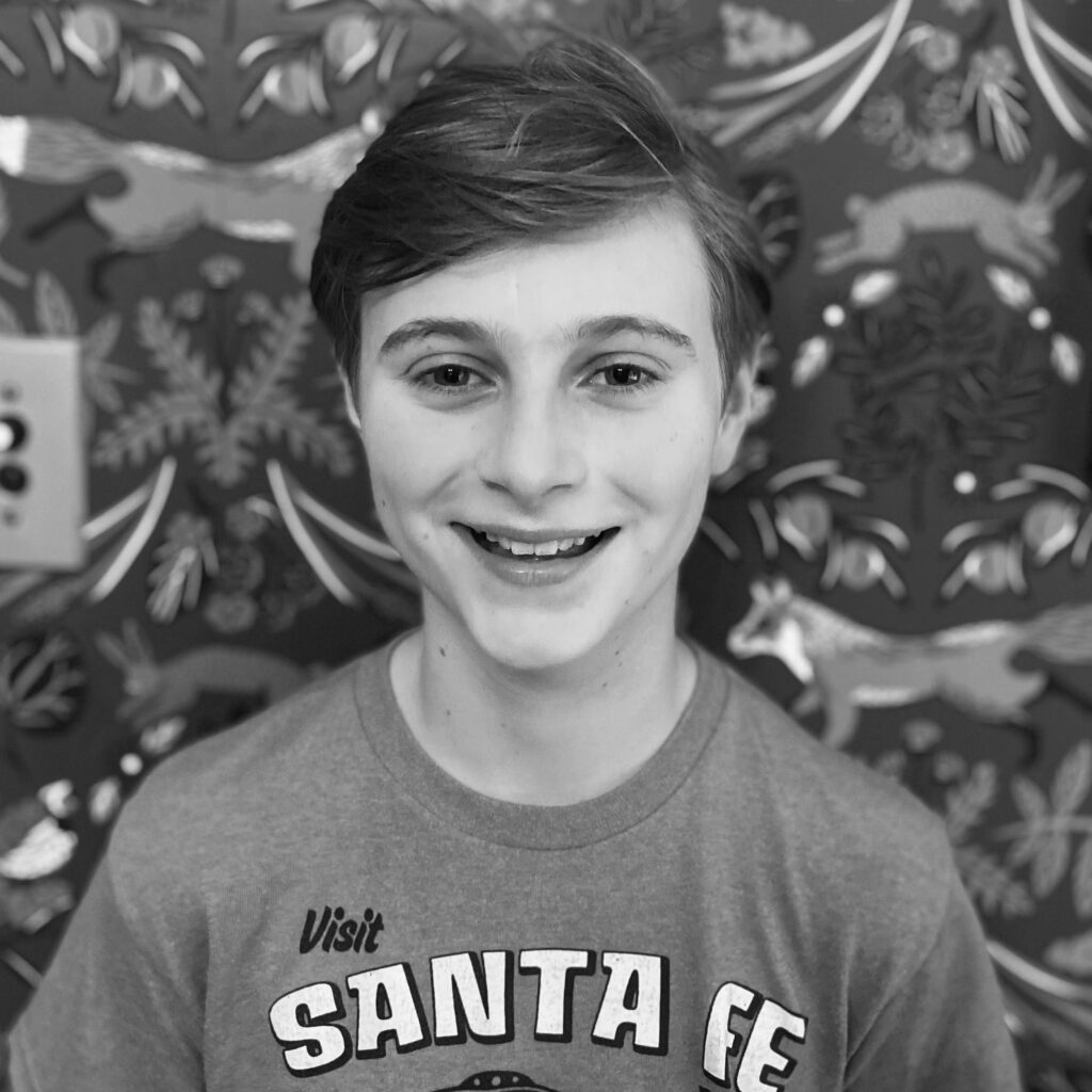 A smiling child with short hair wearing a "Visit Santa Fe" t-shirt stands against a decorative wall with animal and plant motifs. The image is in black and white.