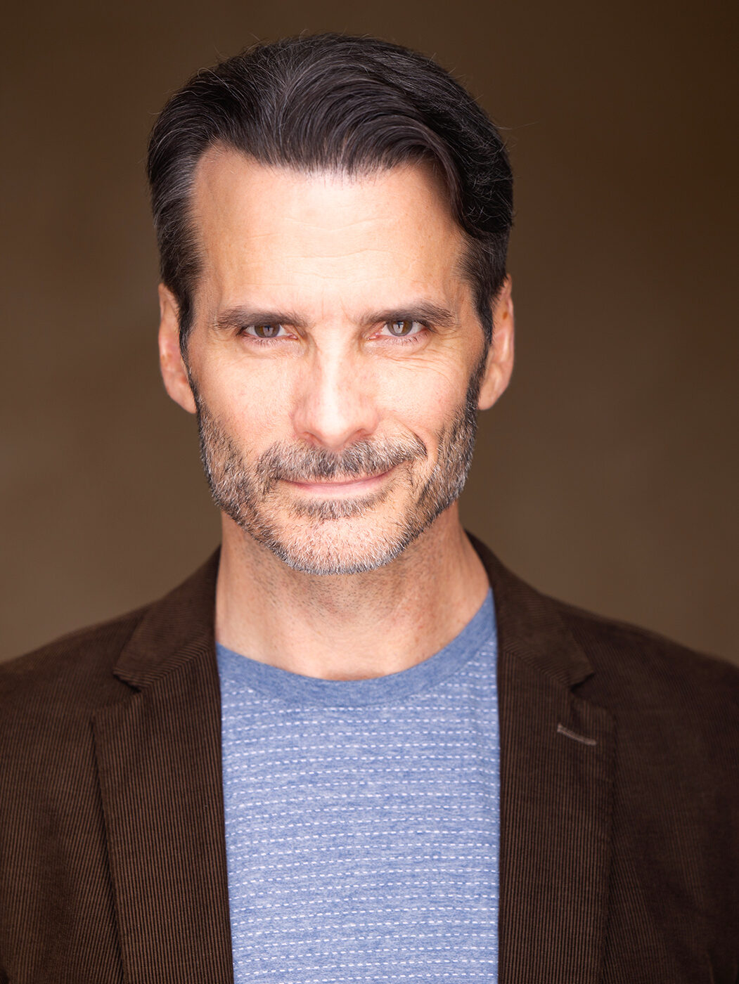 A man with short dark hair and a beard is smiling slightly. He is wearing a brown jacket over a blue textured shirt against a plain brown background.