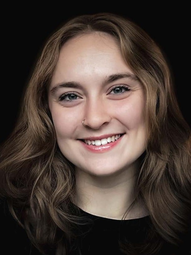 A young woman with wavy brown hair, light skin, and blue eyes smiles at the camera. She is wearing a nose ring and a black top, with a dark, solid background.